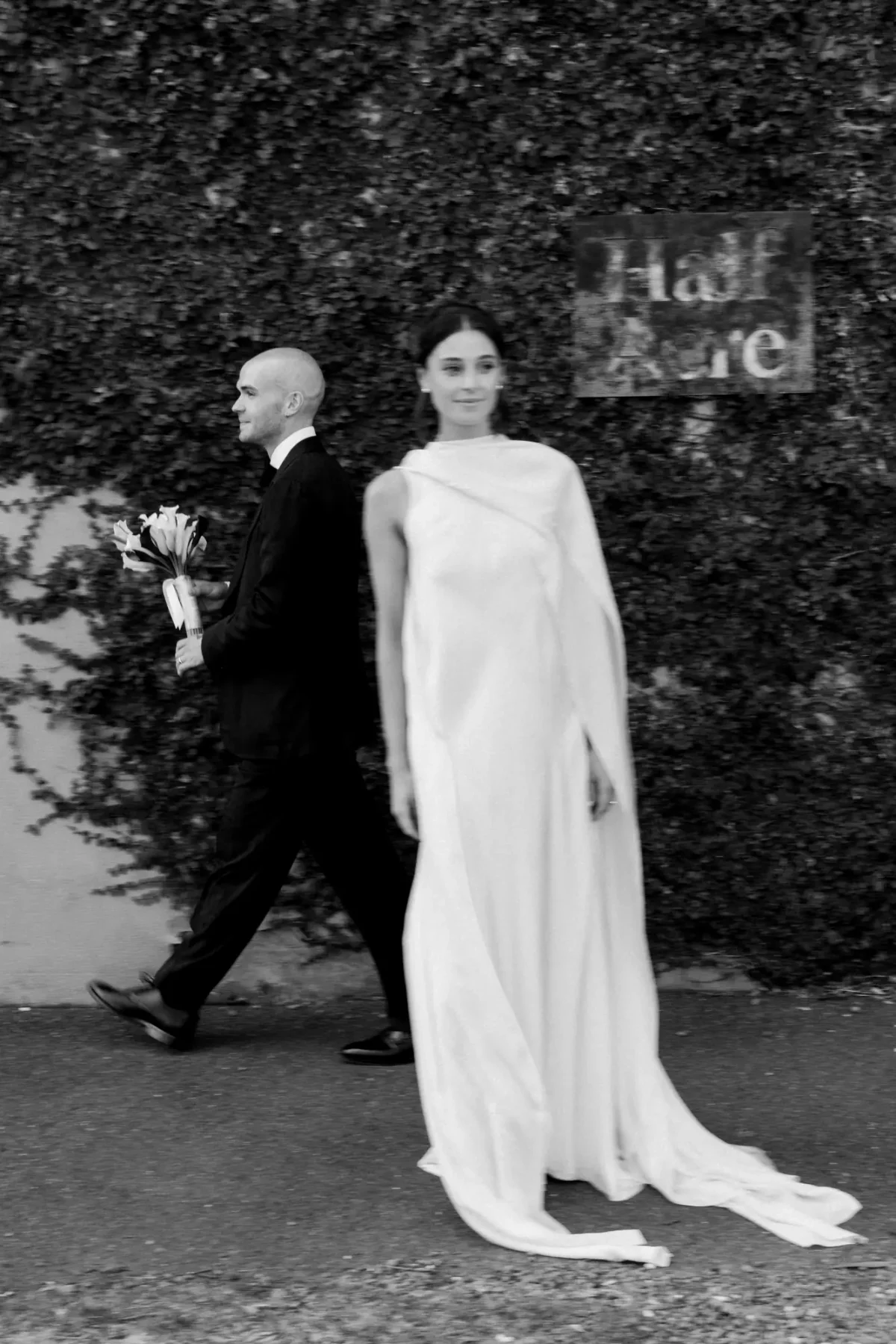 Bride and groom walking past the ivy-covered wall and Half Acre sign