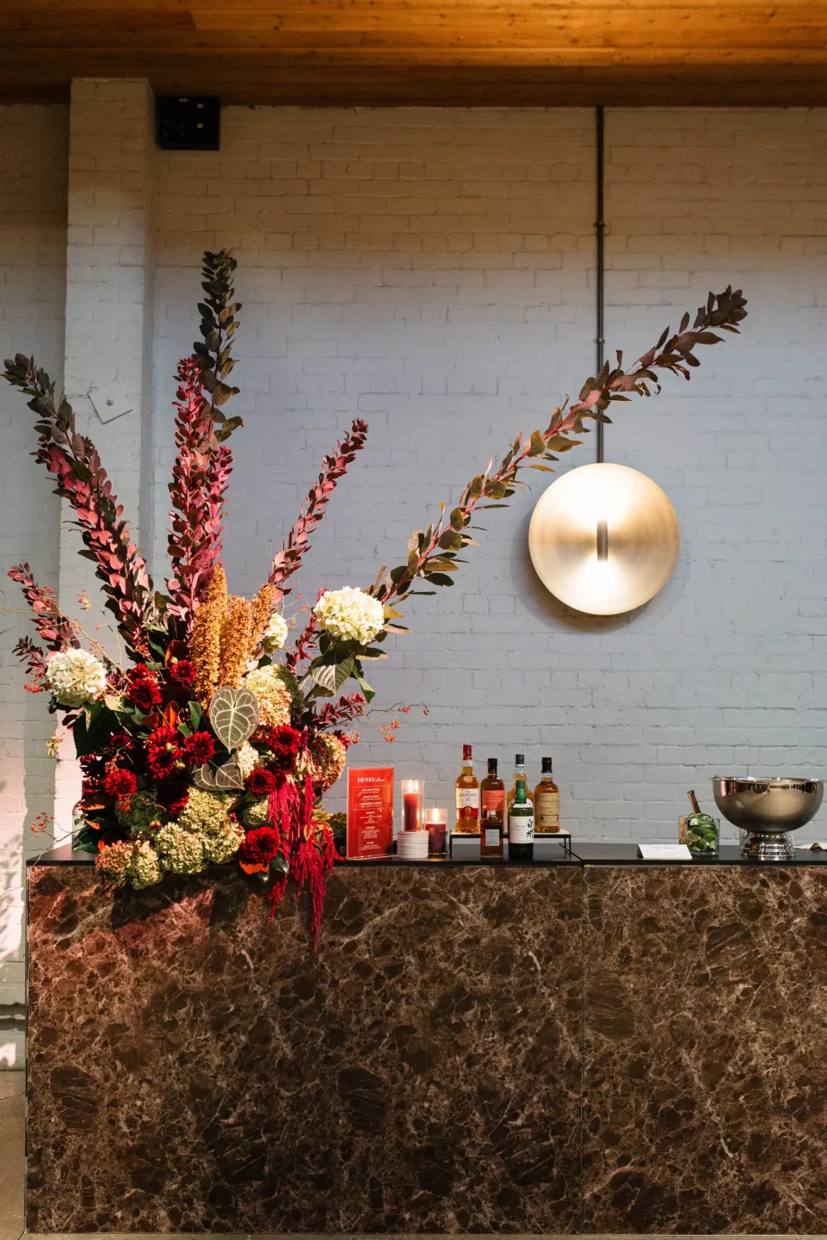 Marble bar styled with a tall red floral arrangement beneath a brass wall light at Half Acre