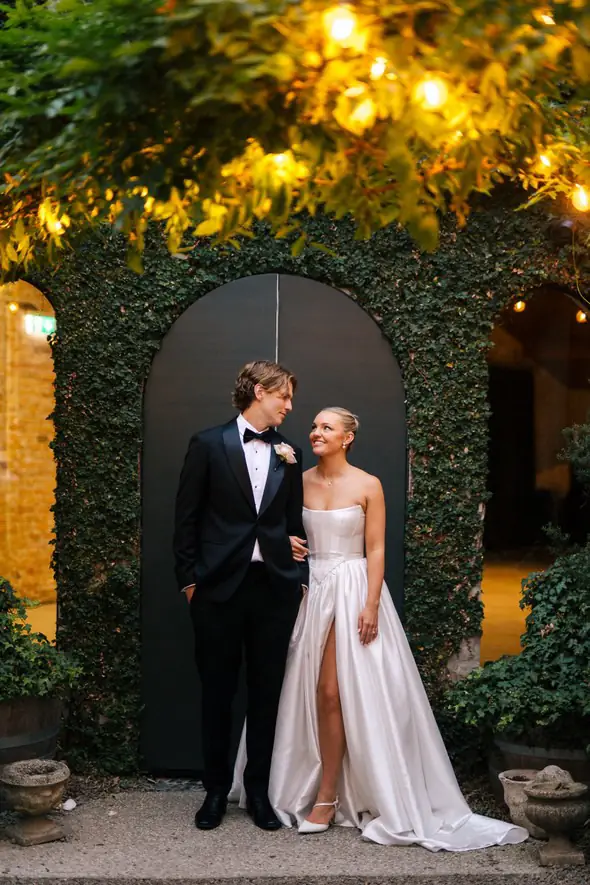 Bride and groom in front of the ivy-framed arched door under festoon lights at Half Acre