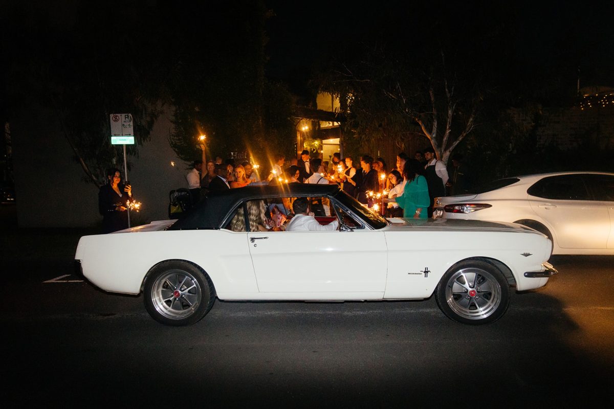 Newlyweds leaving Half Acre at night in a white vintage convertible as guests wave sparklers